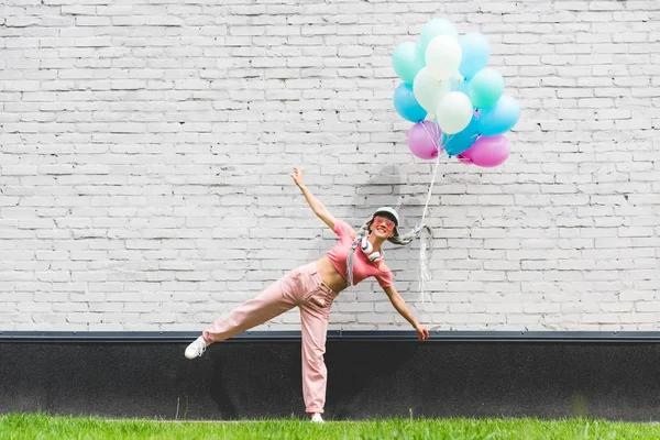 Menina sorridente com balões decorativos e mãos estendidas posando perto da parede de tijolo — Fotografia de Stock