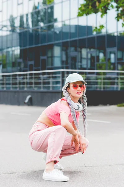 Menina elegante em óculos de sol com fones de ouvido sentado e posando perto do edifício — Fotografia de Stock