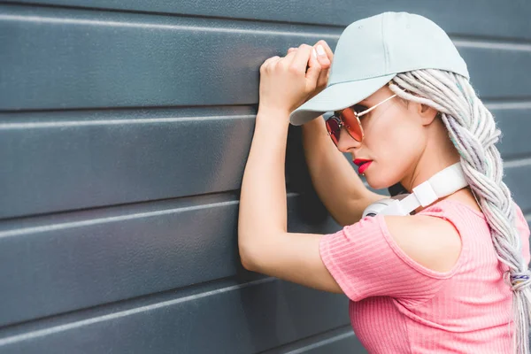 Fashionable girl with dreadlocks and headphones leaning on wall — Stock Photo