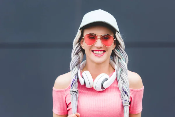 Hermosa chica elegante con rastas y auriculares sonriendo y mirando a la cámara - foto de stock