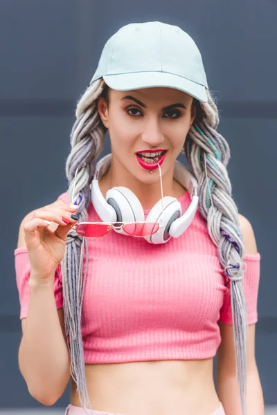 Beautiful stylish girl with dreadlocks and headphones biting sunglasses — Stock Photo