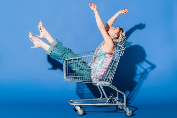 Beautiful stylish girl in shopping cart gesturing on blue — Stock Photo