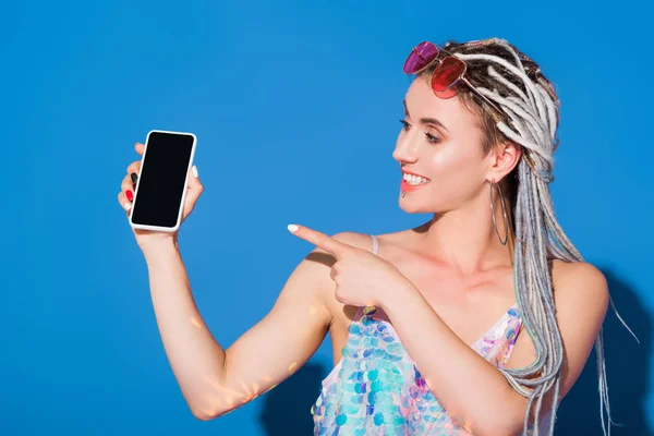 Beautiful stylish girl pointing with finger at smartphone with blank screen on blue — Stock Photo