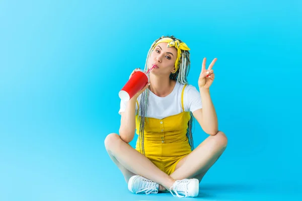 Belle fille avec dreadlocks assis, montrant signe de paix et boire de la soude sur turquoise — Photo de stock