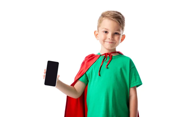 Front view of smiling preschooler kid in red hero cloak showing smartphone with blank screen isolated on white — Stock Photo
