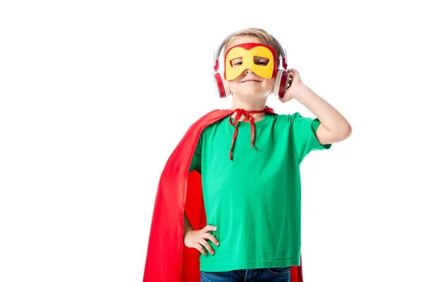 Vue de face de l'enfant préscolaire souriant dans le masque et manteau héros rouge écouter de la musique dans les écouteurs et en regardant la caméra isolée sur blanc — Photo de stock