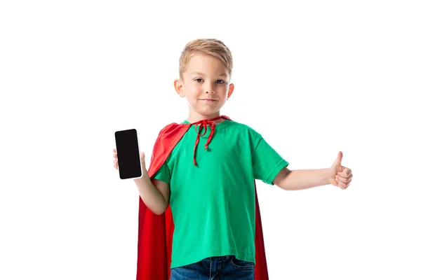 Vista frontal de criança pré-escolar sorridente em capa de herói vermelho mostrando smartphone com tela em branco e mostrando polegar isolado no branco — Fotografia de Stock