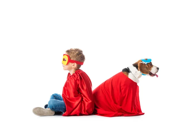 Enfant d'âge préscolaire et chien beagle dans des masques et des sabots de héros rouge assis dos à dos isolé sur blanc — Photo de stock