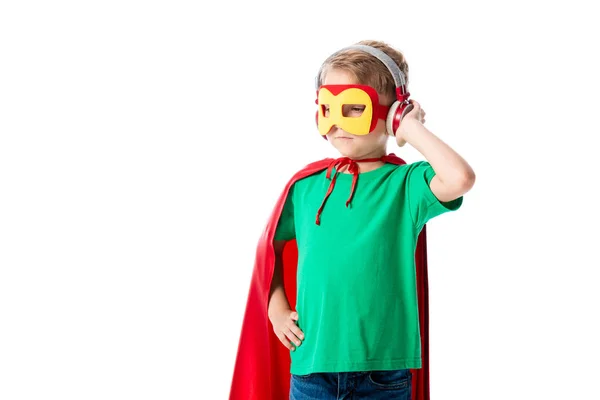 Niño preescolar en capa de héroe rojo y máscara escuchando música en auriculares y mirando hacia otro lado aislado en blanco - foto de stock