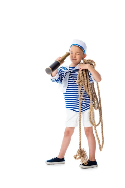 Vista integrale del bambino prescolastico concentrato in costume da marinaio guardando in cannocchiale e tenendo la corda isolata sul bianco — Foto stock