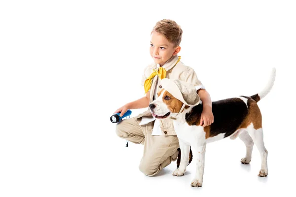 Sonriente explorador niño abrazando lindo perro beagle en sombrero y sosteniendo la linterna en blanco - foto de stock