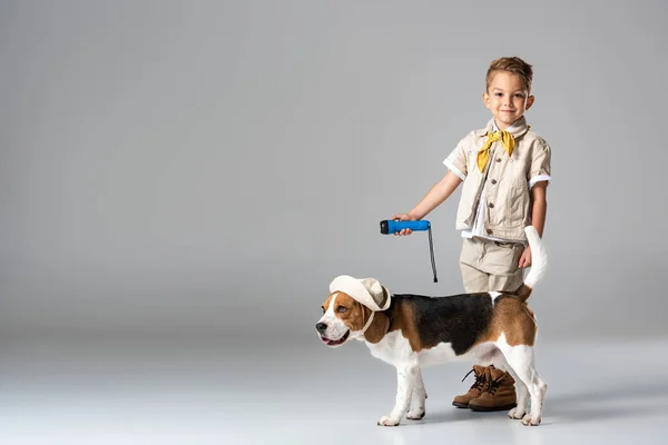 Visão de comprimento total da criança explorador segurando lanterna e cão beagle bonito em chapéu no cinza — Fotografia de Stock