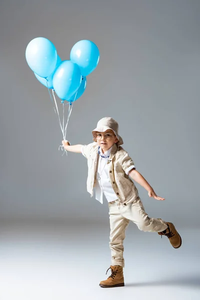 Full length view of explorer child in glasses standing on one leg and holding blue balloons on grey — стоковое фото