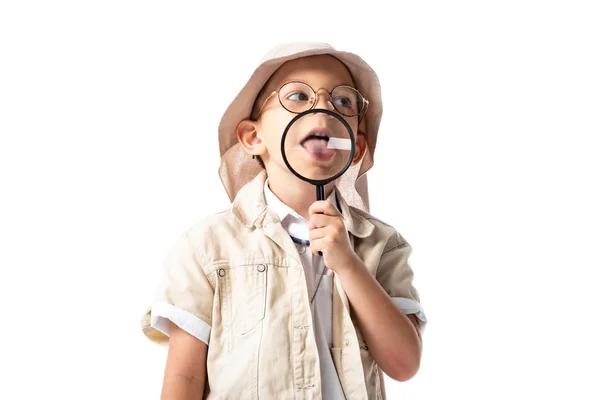 Bambino esploratore in cappello e occhiali tenendo lente d'ingrandimento e sporgente lingua isolata su bianco — Foto stock