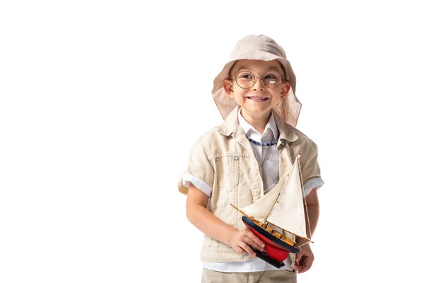 Sonriente explorador niño en gafas y sombrero celebración juguete barco aislado en blanco - foto de stock