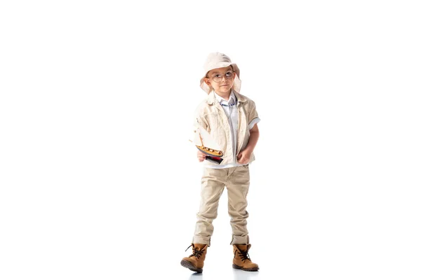 Vue pleine longueur de l'enfant explorateur dans des lunettes et chapeau tenant le bateau jouet sur blanc — Photo de stock