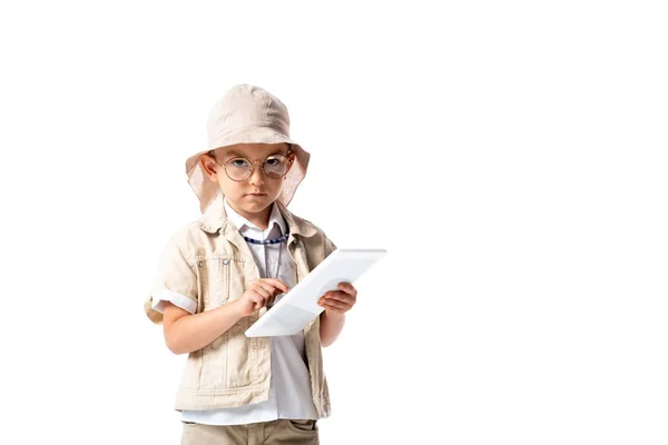 Niño explorador serio en gafas y sombrero sosteniendo tableta digital y mirando a la cámara aislada en blanco - foto de stock