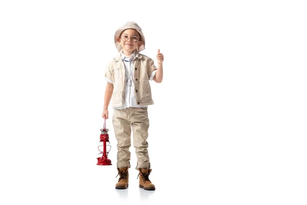 Vue pleine longueur du garçon explorateur souriant dans des lunettes et chapeau tenant lanterne rouge et montrant pouce vers le haut sur blanc — Photo de stock