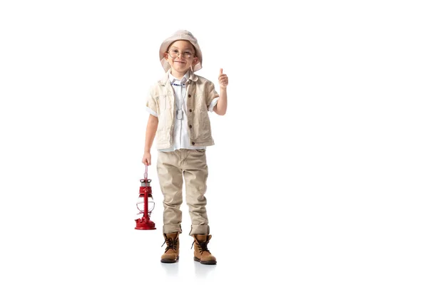 Full length view of smiling explorer boy in glasses and hat holding red lantern and showing thumb up on white — Stock Photo