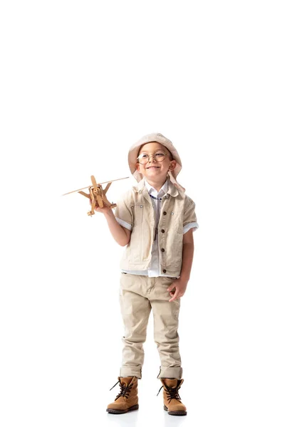 Vue pleine longueur du garçon explorateur souriant dans des lunettes et chapeau tenant avion jouet en bois isolé sur blanc — Photo de stock