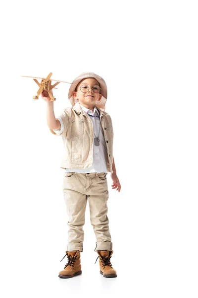 Visão de comprimento total do menino explorador sorridente em óculos e chapéu segurando avião de brinquedo de madeira isolado no branco — Fotografia de Stock