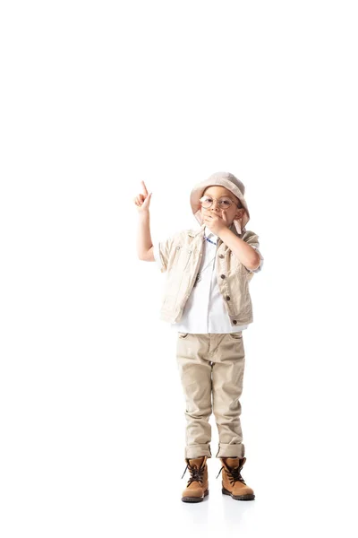 Vista completa de niño explorador sorprendido en sombrero y gafas que muestran signo de idea y la boca cubierta con la mano en blanco - foto de stock