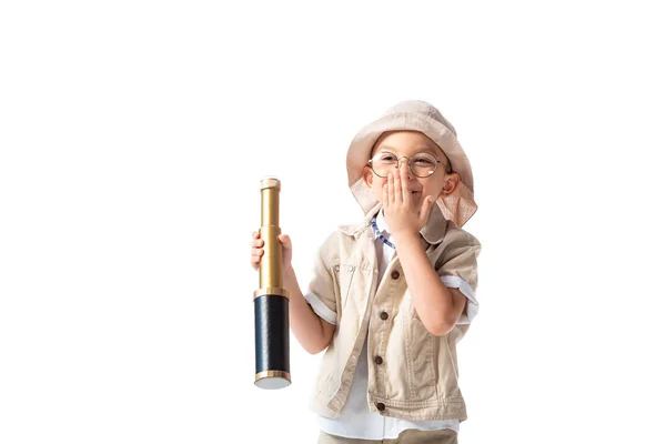 Smiling explorer boy in glasses and hat holding spyglass and covering mouth with hand isolated on white — Stock Photo