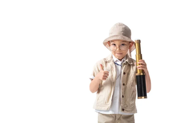 Vista frontal do menino explorador sorridente em óculos e chapéu segurando spyglass e mostrando polegar para cima isolado no branco — Fotografia de Stock