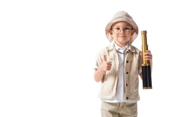 Vista frontale del ragazzo esploratore sorridente in occhiali e cappello tenendo spyglass e mostrando pollice in alto isolato su bianco — Foto stock