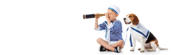 Panoramic shot of smiling boy in sailor suit looking through spyglass and beagle dog on white — Stock Photo