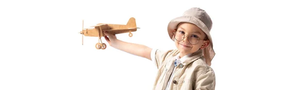 Панорамный снимок улыбающегося ребенка-исследователя в очках и шляпе, держащего деревянный игрушечный самолет, изолированный на белом — стоковое фото