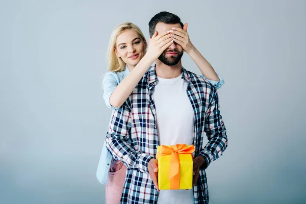 Beautiful girl Covering Eyes of man holding present on grey — Stock Photo