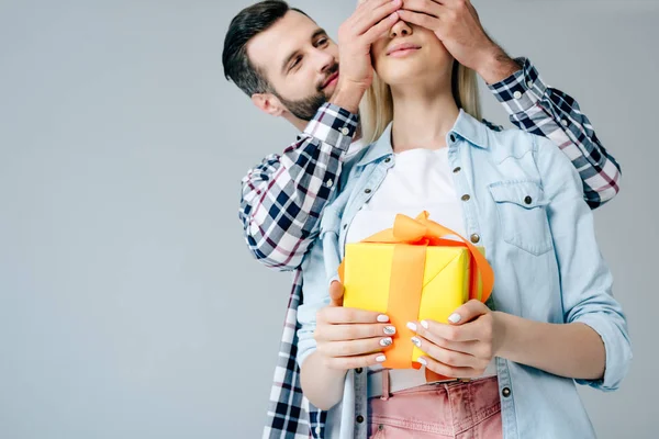 Bonito homem cobertura olhos de menina segurando presente isolado no cinza — Fotografia de Stock