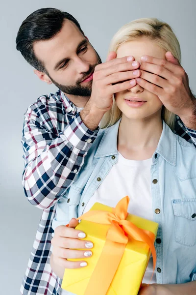 Чоловік покриває очі молодої жінки подарунком ізольовано на сірому — стокове фото