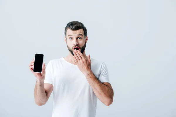 Hombre sorprendido cubriendo la boca y mostrando teléfono inteligente con pantalla en blanco aislado en gris - foto de stock