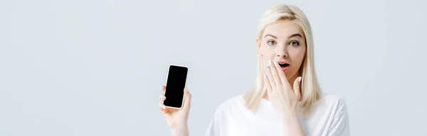 Plano panorámico de hermosa chica impactada mostrando teléfono inteligente con pantalla en blanco aislado en gris - foto de stock