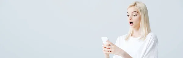 Панорамный снимок красивой потрясенной блондинки с помощью смартфона, изолированного на сером — стоковое фото
