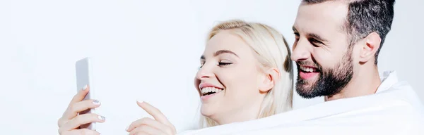 Panoramic shot of man and beautiful girl covered in blanket using smartphone on white — Stock Photo