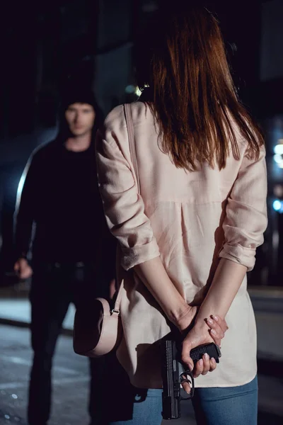 Vista trasera de la mujer escondiendo el arma detrás de la espalda cerca de ladrón en la noche - foto de stock