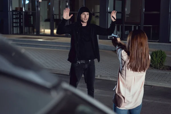 Mujer apuntando pistola al ladrón con las manos en el aire por la noche en el estacionamiento - foto de stock