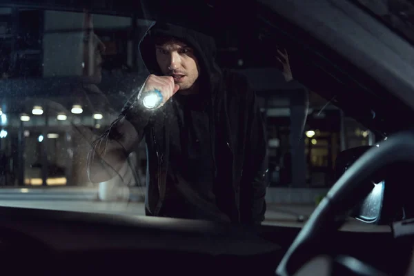 Thief in black hoodie looking into car with flashlight — Stock Photo