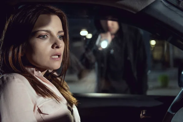 Вор с фонариком заглядывает в машину с напуганной женщиной — стоковое фото