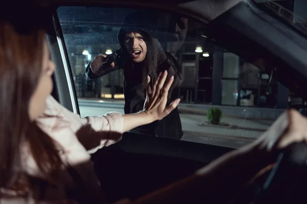 Ladrón enojado apuntando arma a la mujer sentada en el coche por la noche - foto de stock