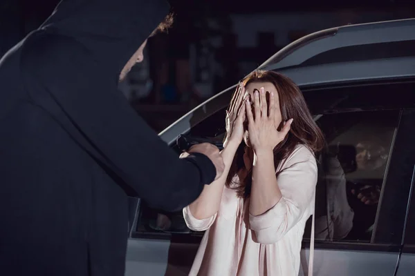 Ladrón atacando mujer cubriendo la cara cerca de coche por la noche - foto de stock