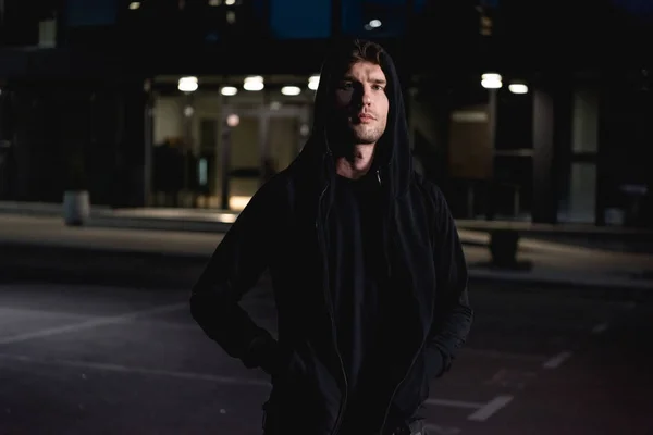 Thief in black hoodie looking at camera in parking lot at night — Stock Photo