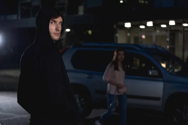 Enfoque selectivo de ladrón mirando a la cámara cerca de mujer caminando por la noche - foto de stock