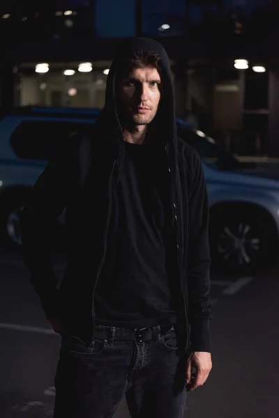 Ladrão com capuz preto olhando para a câmera no estacionamento à noite — Fotografia de Stock