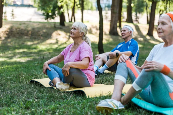 Foco seletivo de mulheres aposentadas com olhos fechados sentados com pernas cruzadas perto do homem sênior no parque — Fotografia de Stock