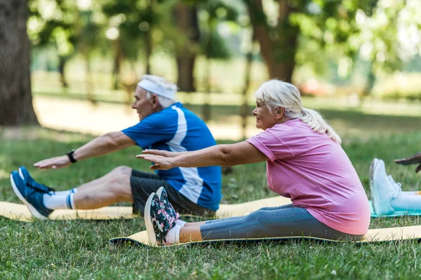 Enfoque selectivo de la mujer mayor con el pelo gris y el hombre haciendo ejercicio en fintess esteras en la hierba - foto de stock