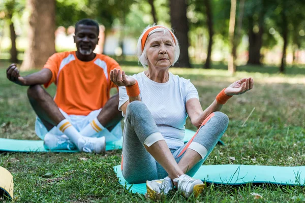Selektiver Fokus einer Rentnerin und eines afrikanisch-amerikanischen Mannes mit geschlossenen Augen, die mit gekreuzten Beinen im Park sitzen — Stockfoto